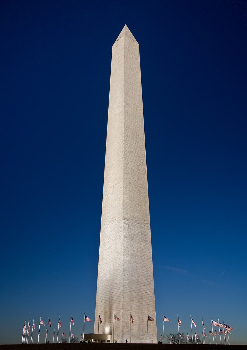 El Monumento a Washington elevándose alto contra un cielo azul intenso al anochecer, rodeado de banderas americanas